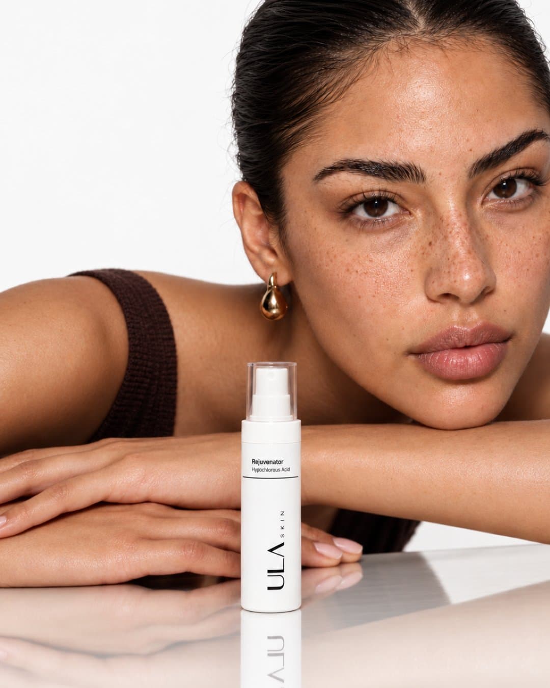 Model resting her chin on folded arms beside a skincare spray bottle on a mirrored table