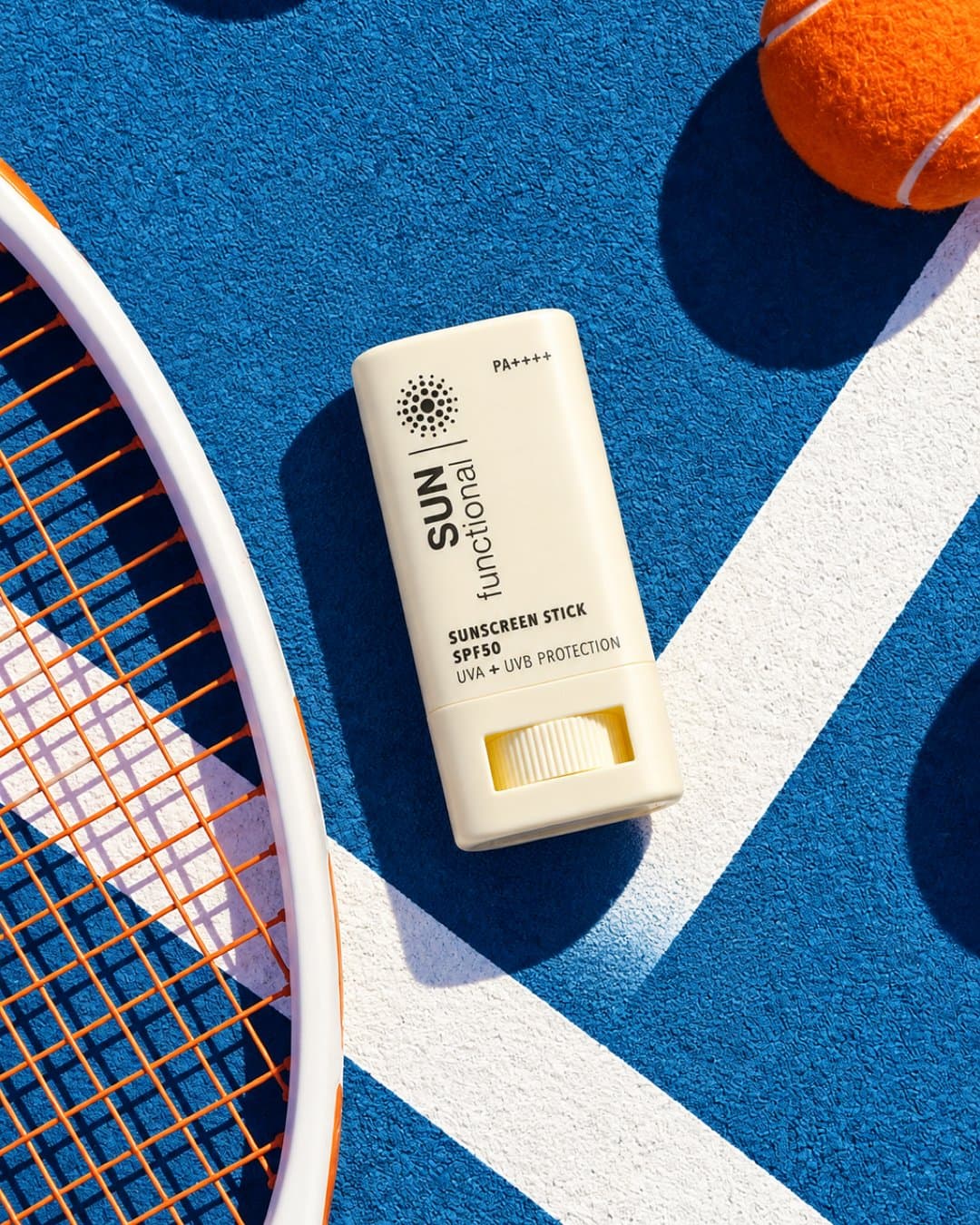 Sunscreen stick on a blue tennis court beside a racket and ball, shot from above