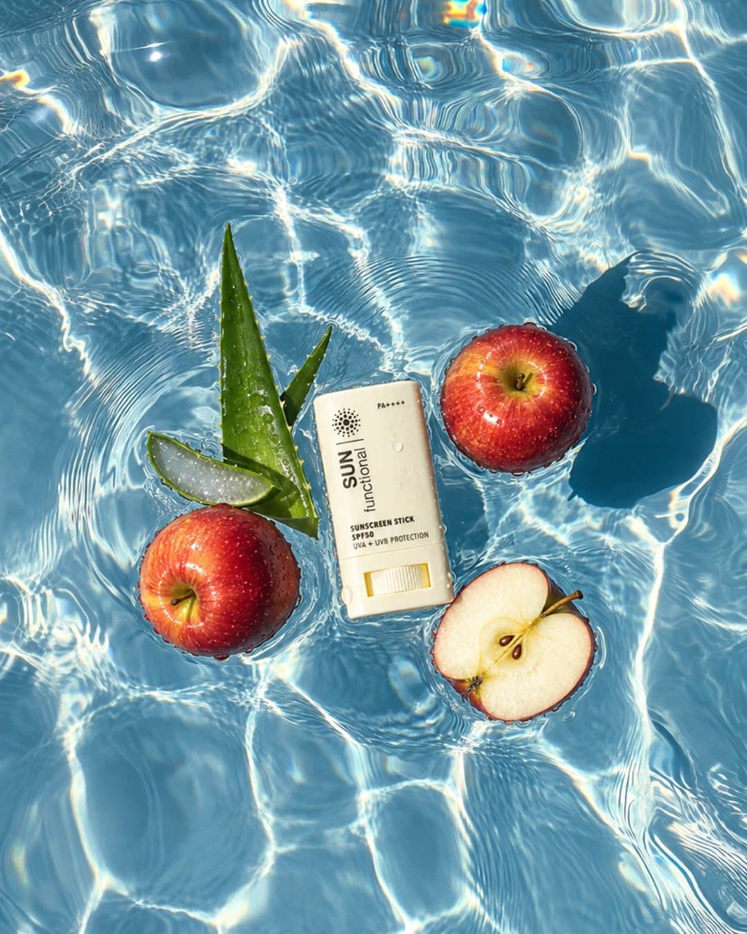 Sunscreen stick floating in a bright blue pool with apple halves and aloe, shot from above