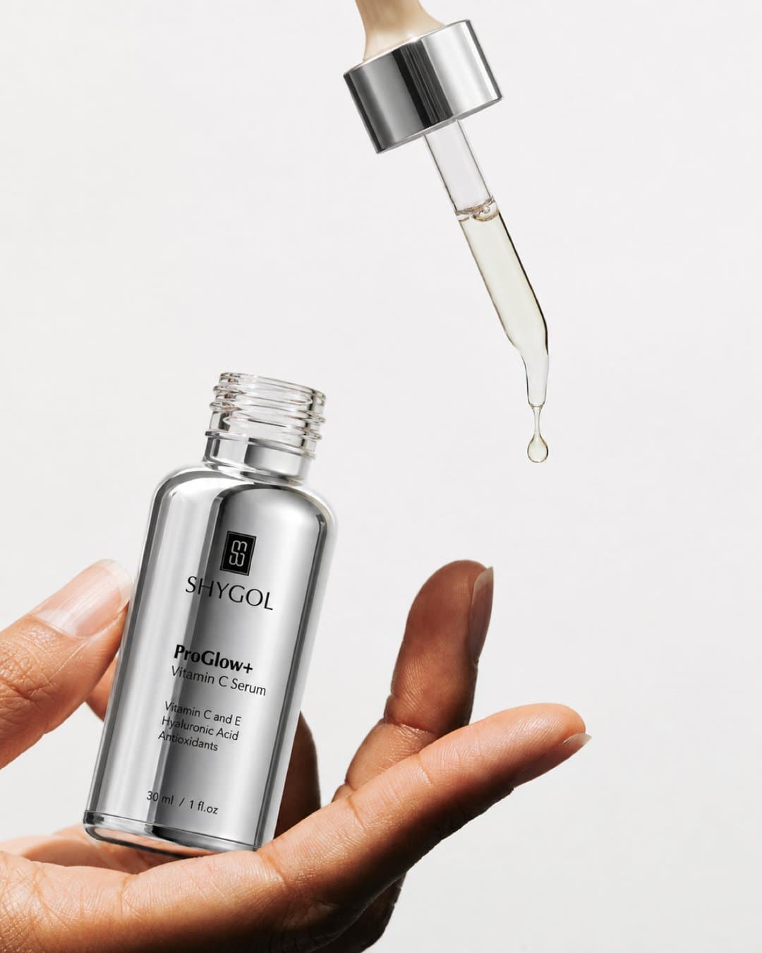 Silver ProGlow+ serum bottle held in an open hand while its dropper releases a drop of serum