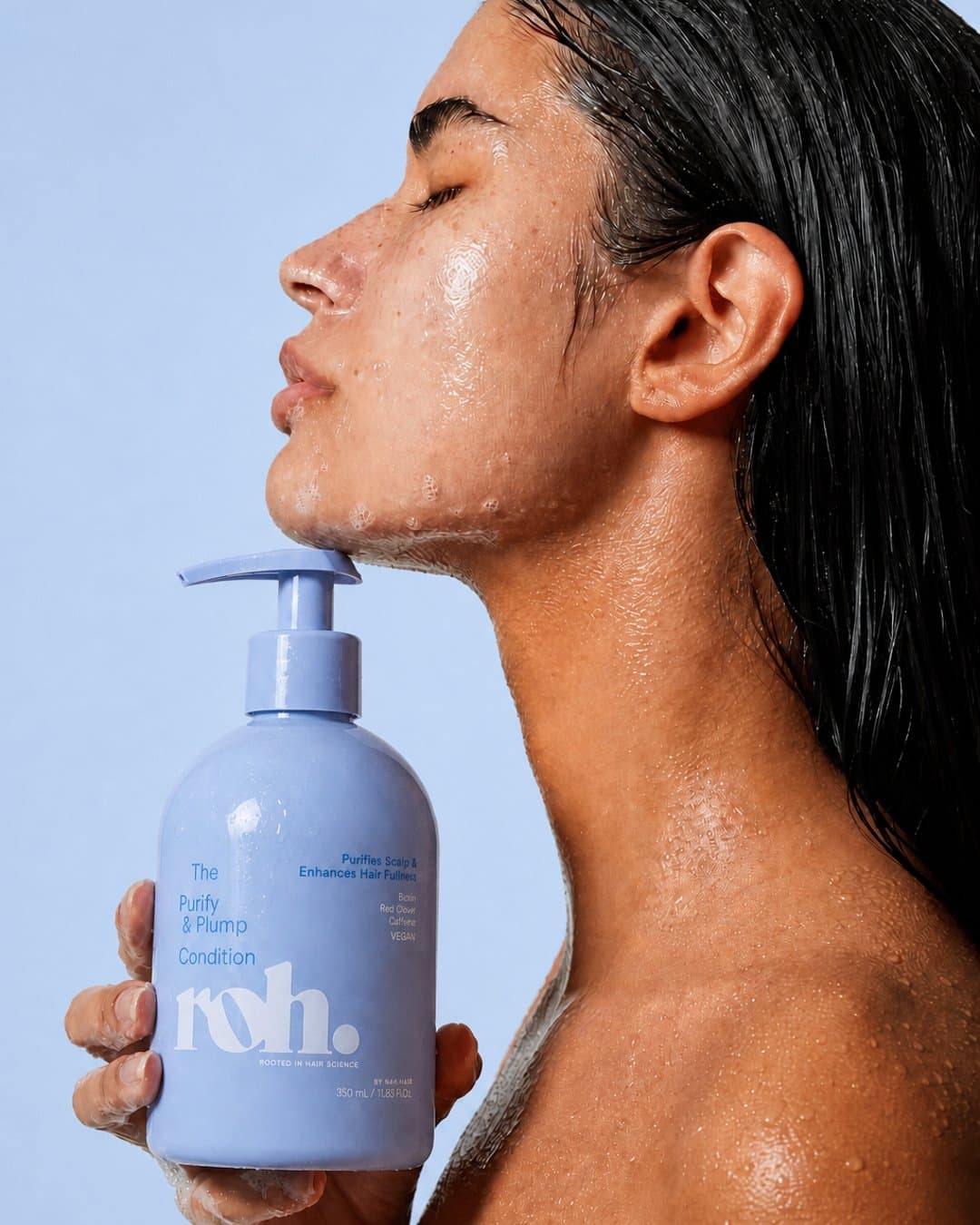 Model in profile with water on her skin, holding a ROH pump bottle at her chin against a blue wall
