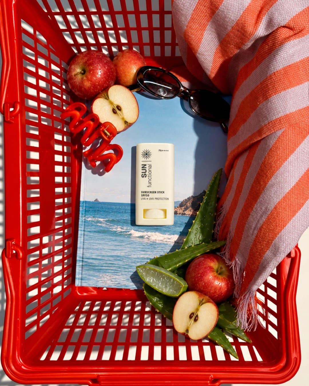 Sunscreen stick in a red shopping basket with apples, aloe and a striped towel above a sea view