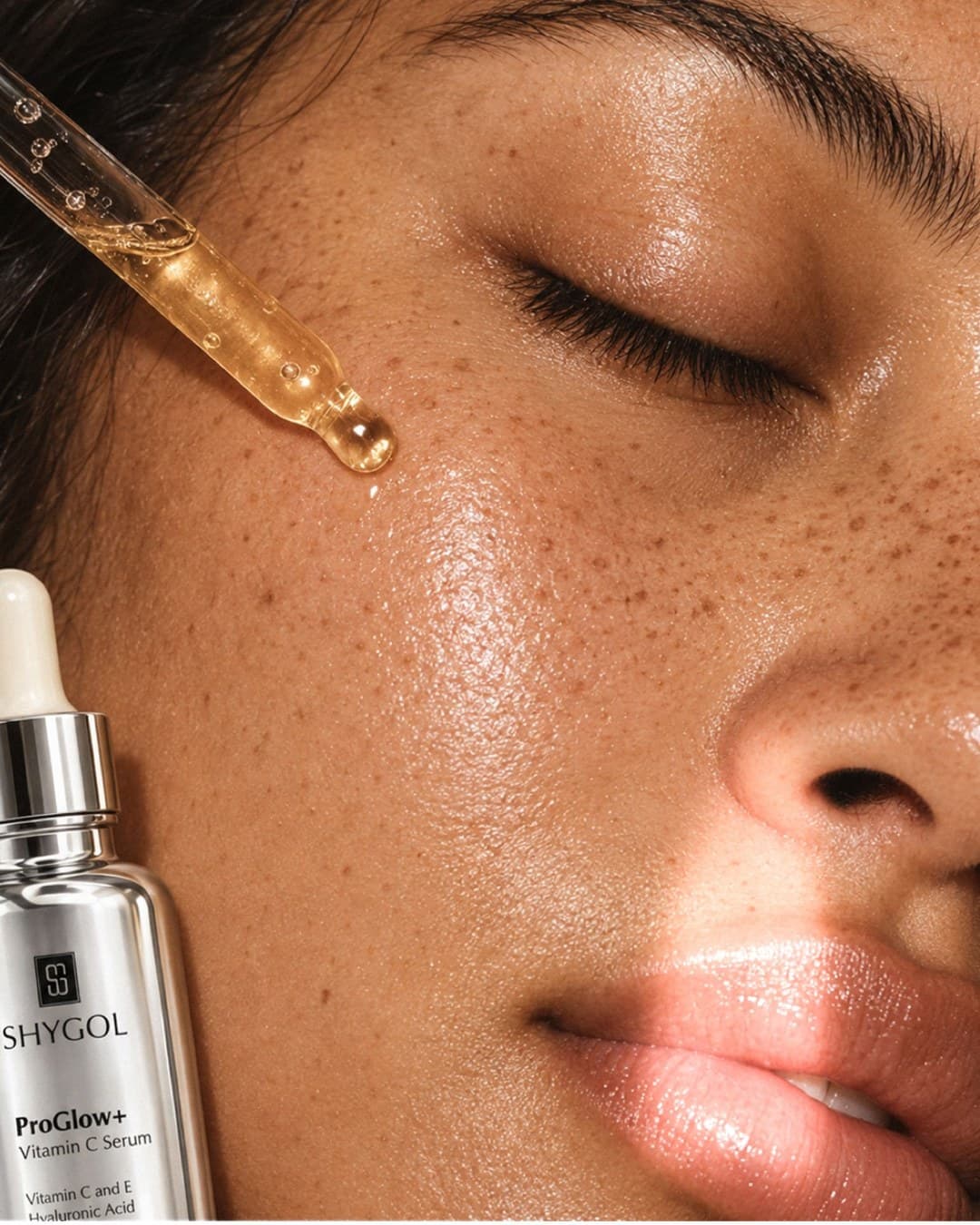 Close-up of a serum dropper releasing a drop onto freckled skin beside a ProGlow+ bottle