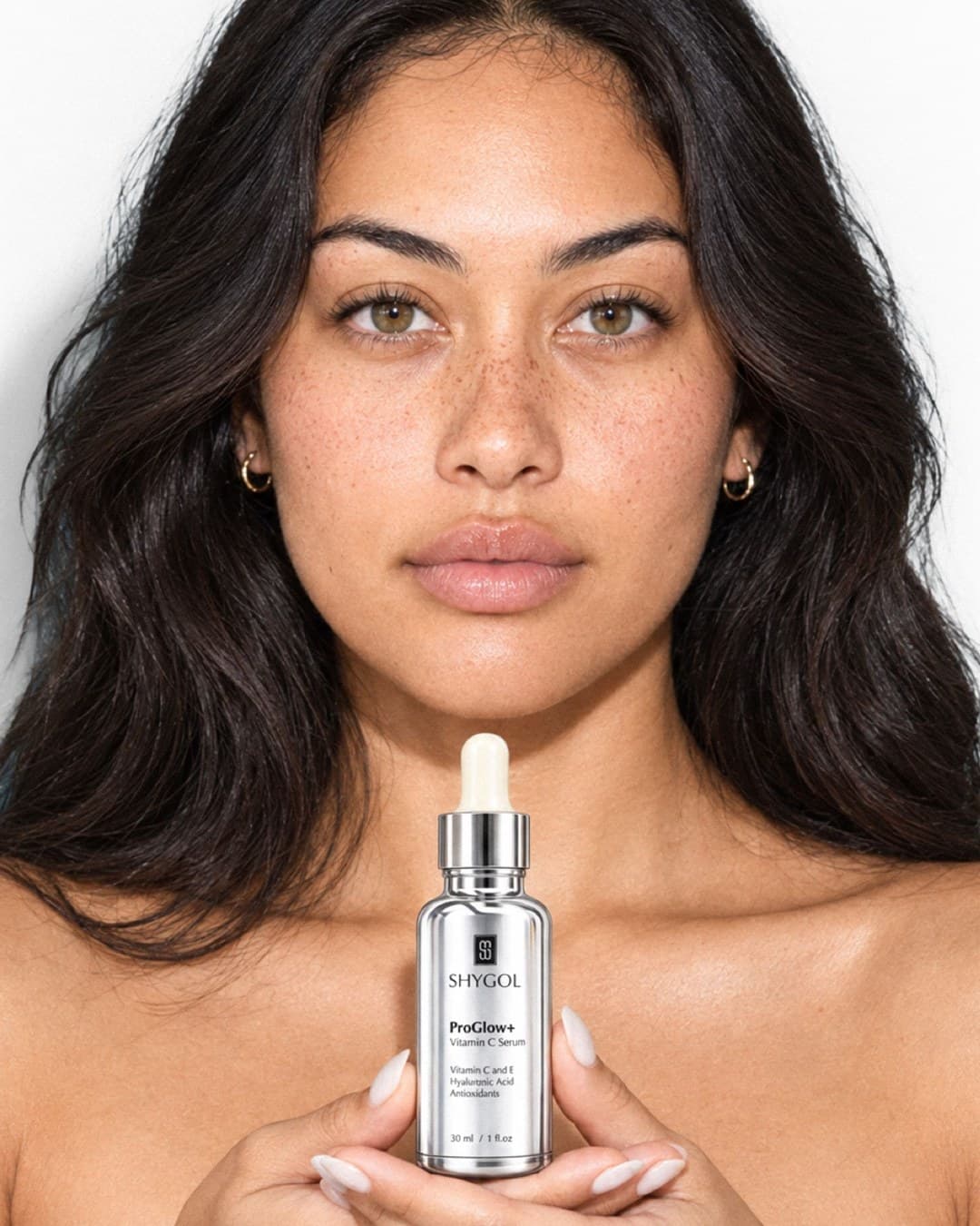 Front-on portrait of a freckled model holding a Shygol ProGlow+ serum bottle under her chin with both hands
