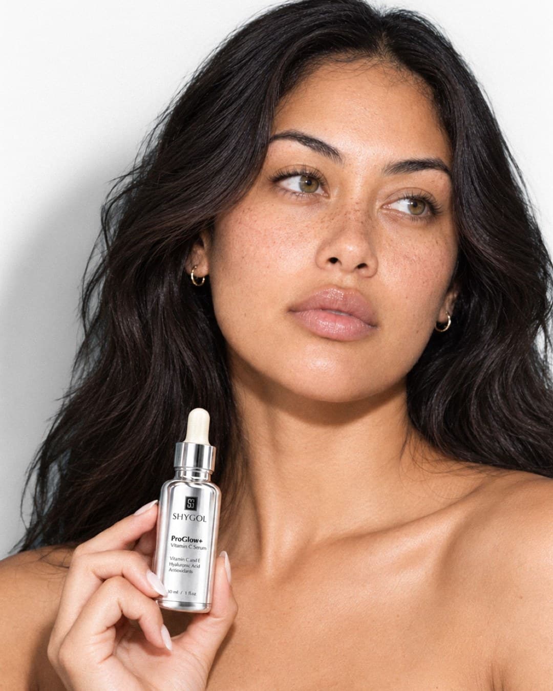 Model looking off to the side and holding a Shygol ProGlow+ serum bottle at her shoulder against a white wall