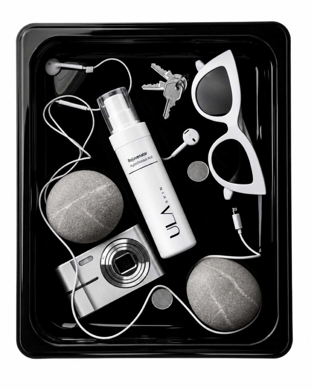 Black and white flat lay of a ULA Rejuvenator spray bottle in a black tray with keys, sunglasses, a camera and earphones