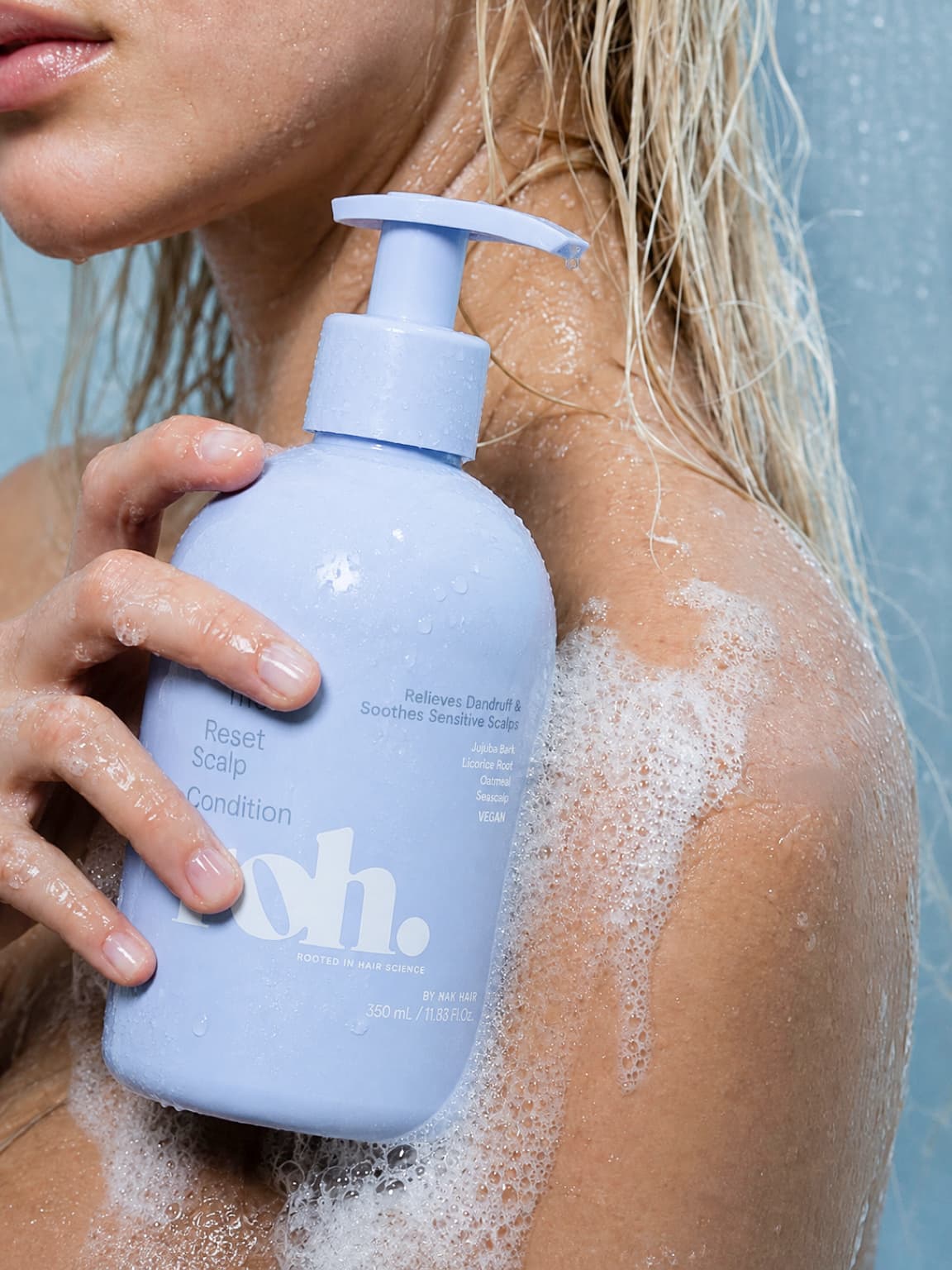 Close crop of a model holding a periwinkle condition bottle to a soapy, wet shoulder in the shower