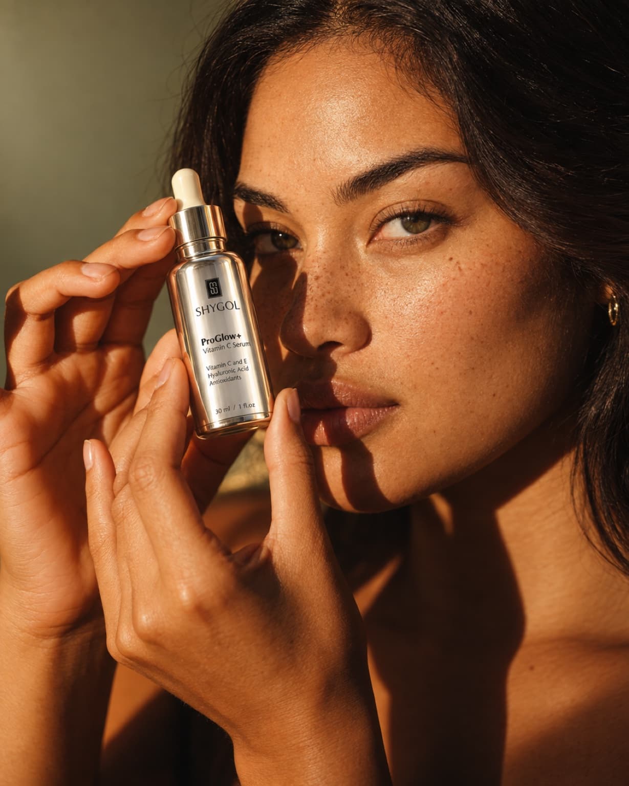 Close beauty shot of a model holding a ProGlow+ Vitamin C Serum bottle beside her cheek in hard side light, freckles and skin texture visible