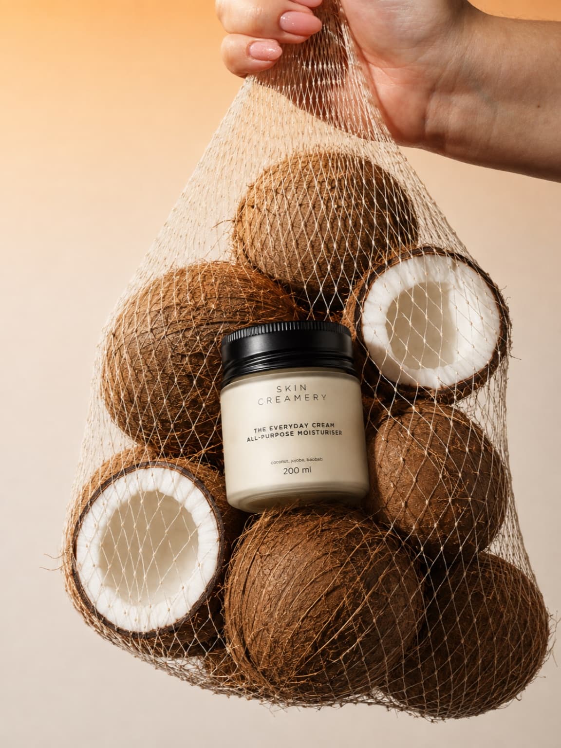 Hand holding a net bag of cracked coconuts with The Everyday Cream jar nested among them, warm backlight