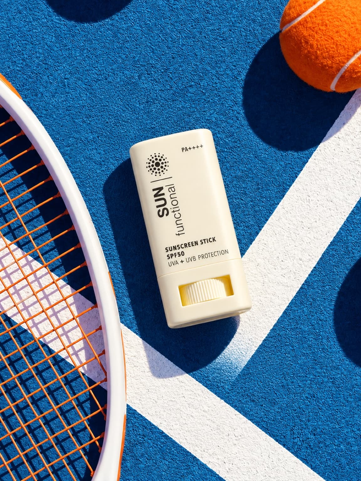 Overhead flat lay of a SUN functional sunscreen stick beside a tennis racket and ball on a hard court